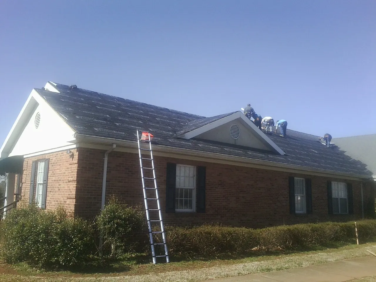 Crew tearing shingles off a brick ranch roof with a ladder set — Emergency Roof Repair work by our crew in Lindsay